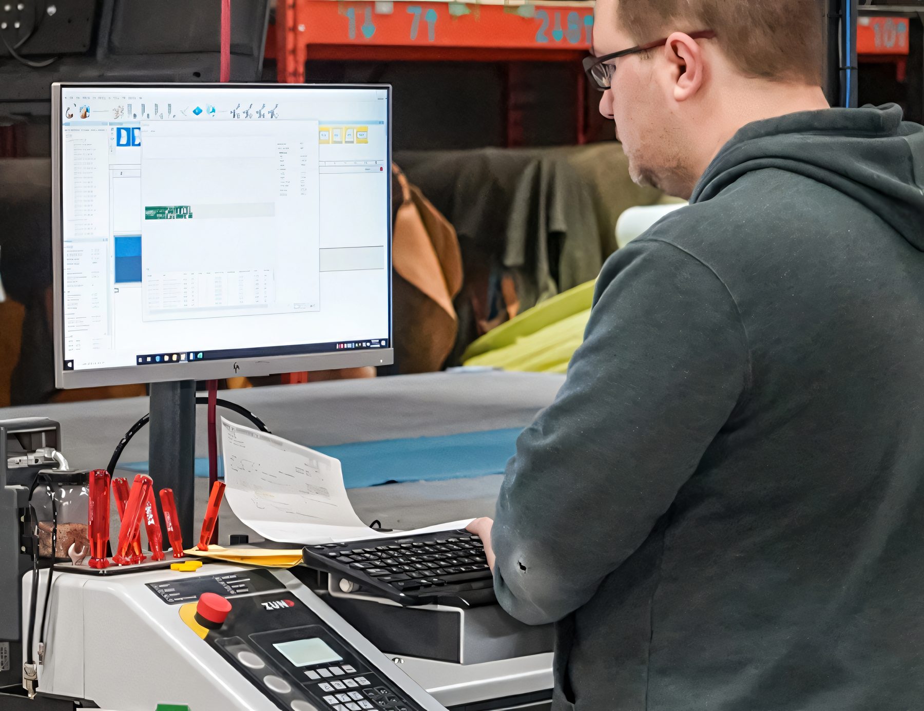 Devant son écran, le programmeur de la table de découpe numérique ZUND prépare un plan de coupe, les rouleaux de tissu derrière lui