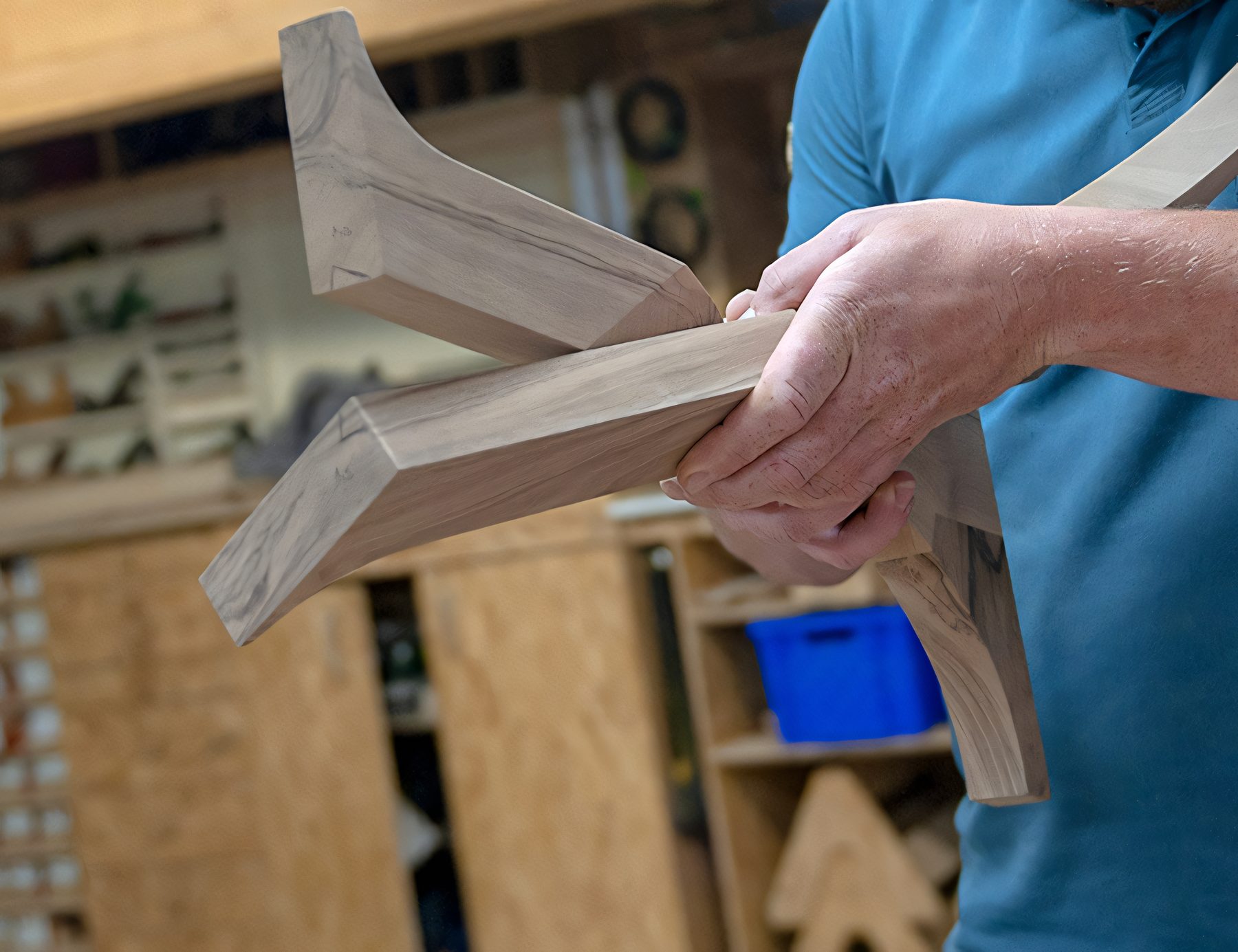 Un ébéniste tient un piètement de bois massif fraîchement assemblé, l'atelier et ses étagères de gabarits derrière lui