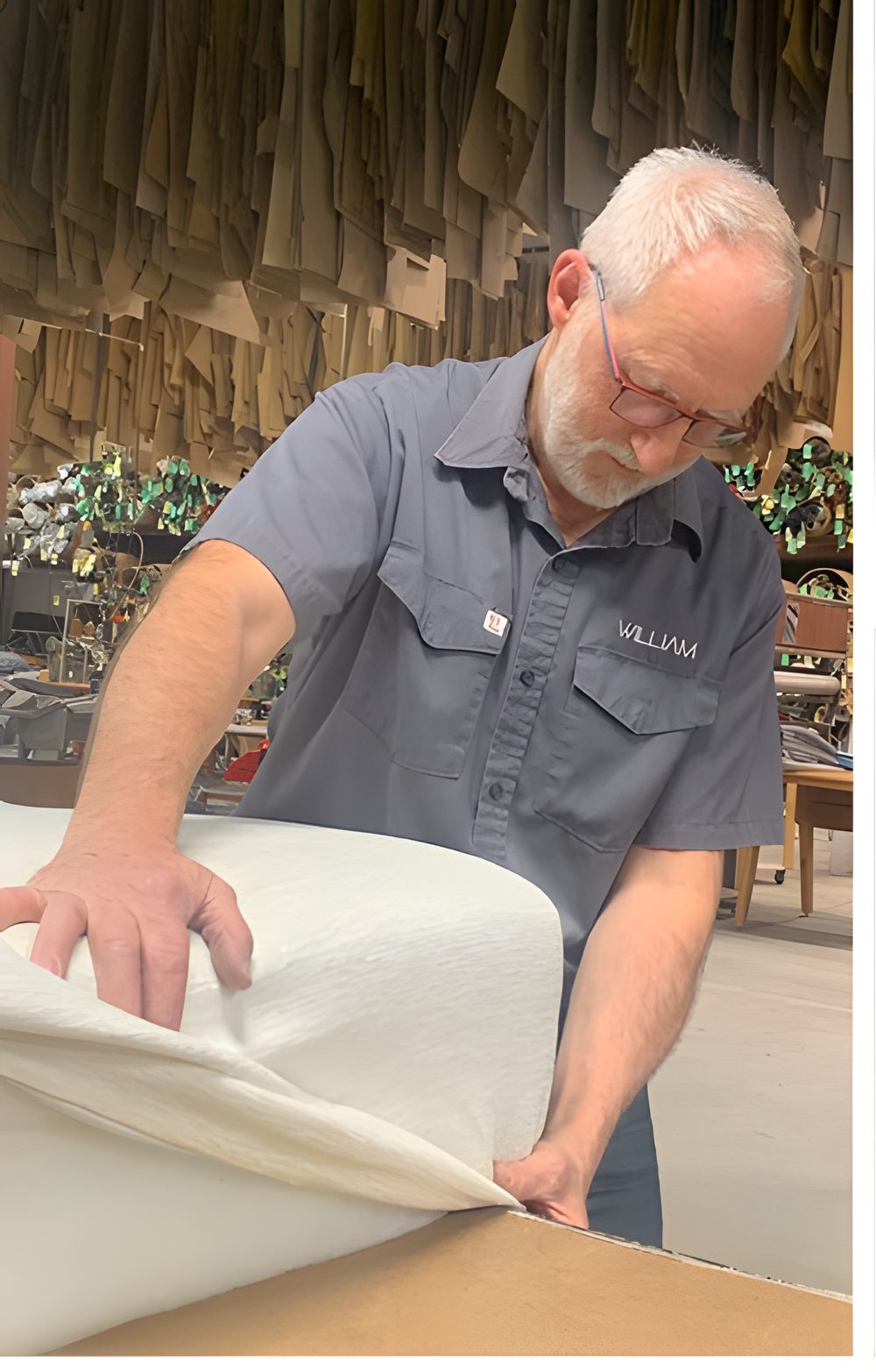Un rembourreur de William, chemise grise brodée du logo, pose une ouate blanche sur un coussin dans l'atelier, des patrons suspendus derrière lui
